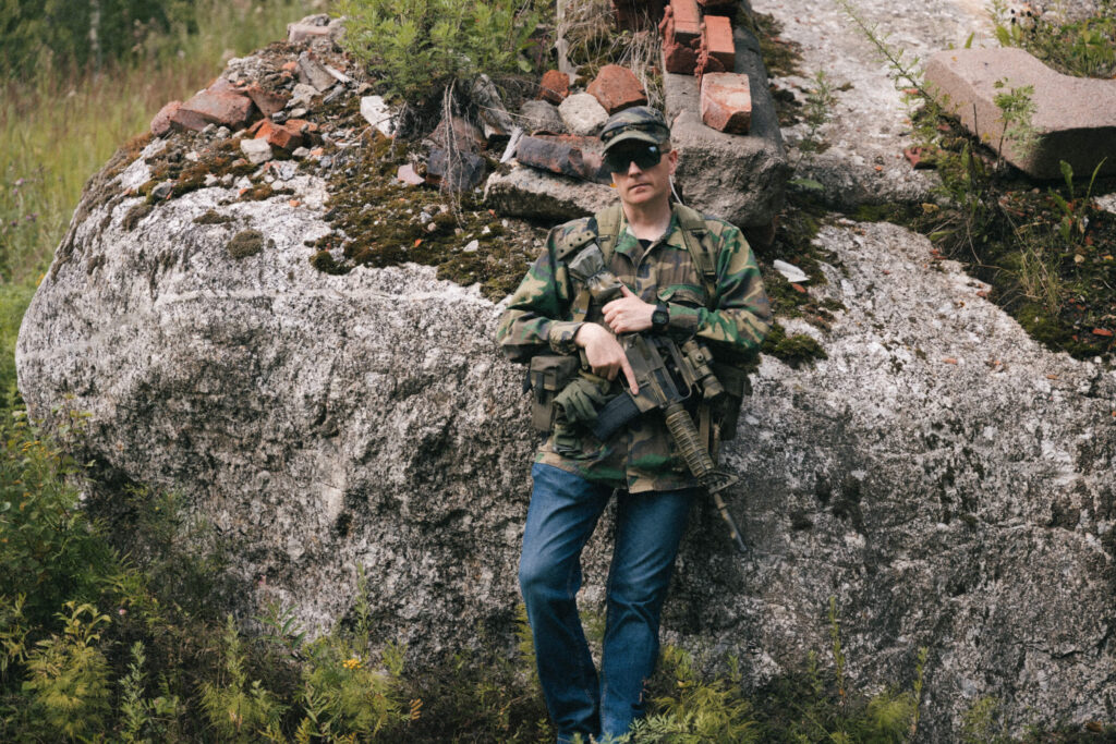 A person leaning on a large rock in camouflage jacket and blue jeans. He is holding the Green Diamond Carbine.